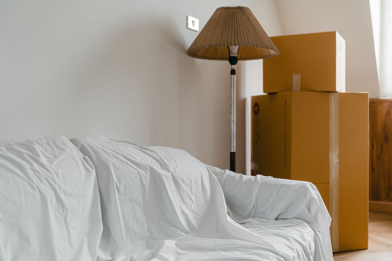who-we-are Covered with sheet sofa and floor lamp and packed cardboard boxes in light spacious room of new contemporary house